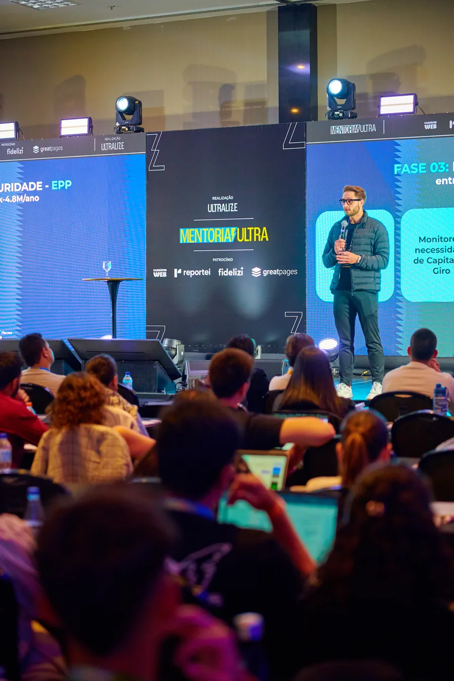 Palestrante no palco da Mentoria Ultra, com a plateia de laptops abertos em primeiro plano.