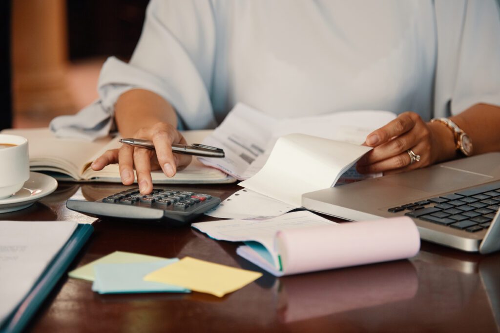 reconciliação bancária: passos para evitar erros e garantir precisão nas finanças 1 mulher utilizando calculadora sobre uma mesa com diversos papéis, planilhas e um computador, ilustrando a rotina de reconciliação bancária em um ambiente de trabalho.