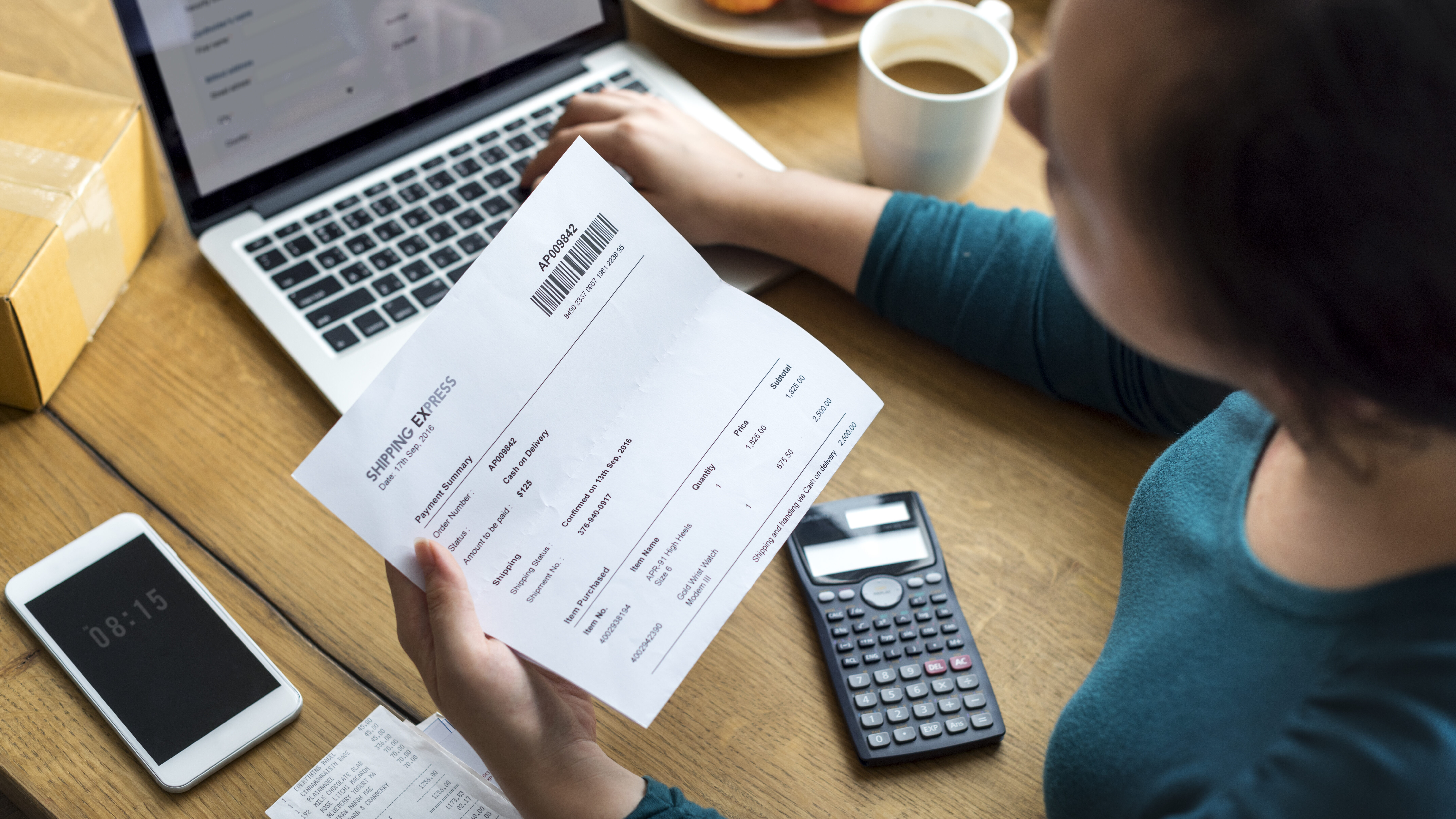 gerenciamento de fluxo de caixa: dicas para manter a saúde financeira da sua empresa 1 mulher vista de cima analisando um papel de conta enquanto insere ou confere dados no computador, representando o gerenciamento de fluxo de caixa em um ambiente empresarial.